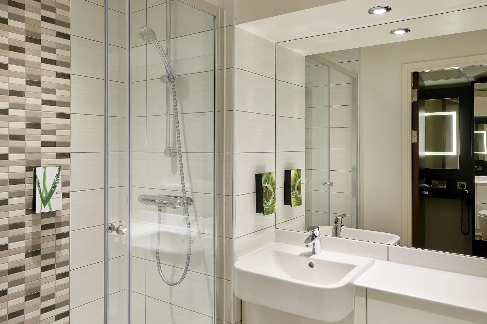 A bathroom at Premier Inn London County Hall with a glass-walled walk-in shower, white tiles with a mosaic strip and a basin under a wide mirror