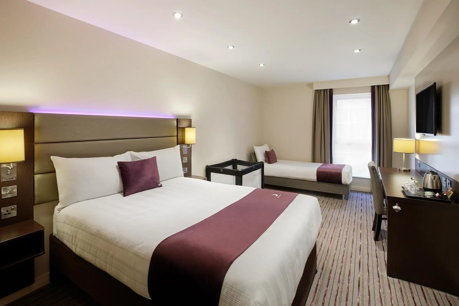 A family room at Premier Inn London County Hall with a double bed, a single bed under the window, a travel cot and a desk with a kettle