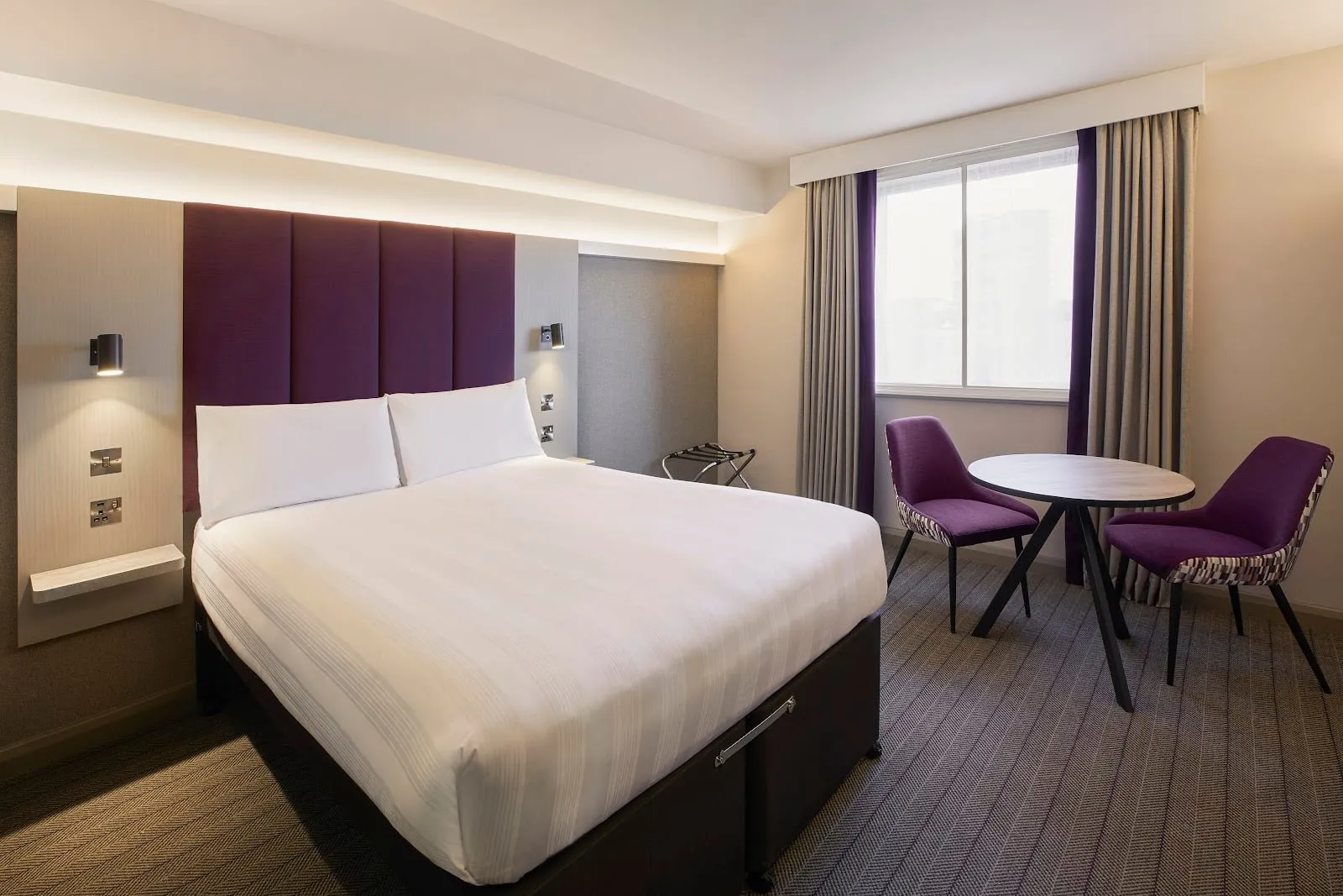 A double room at Premier Inn London Greenwich with a purple headboard and a table and two chairs by the window
