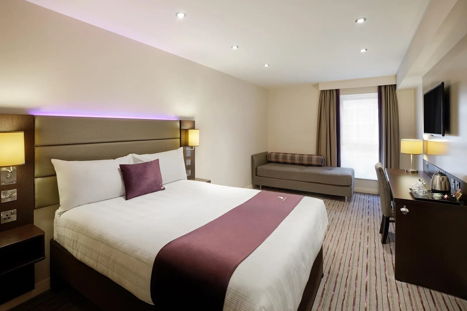 A double room at Premier Inn London Harrow with a padded headboard lit purple from above, a grey chaise longue under the window and a desk with a kettle tray below a wall-mounted TV