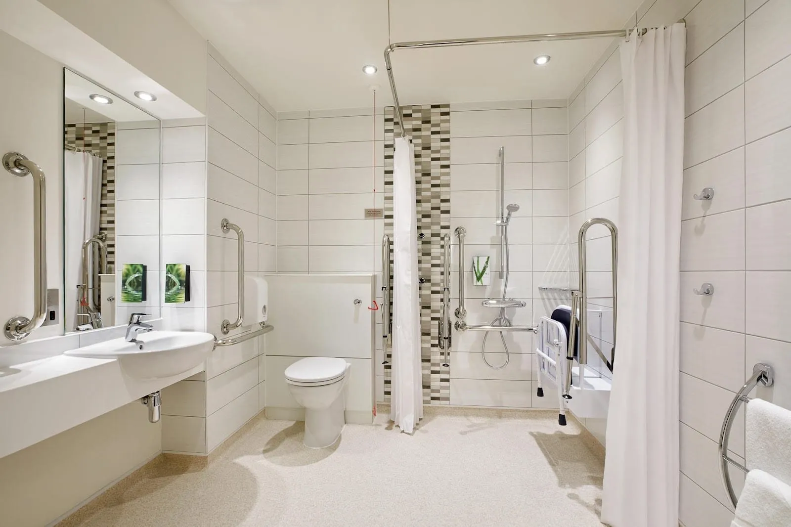 An accessible Premier Inn bathroom with a level-entry shower behind a curtain, a fold-down shower seat, grab rails beside the toilet and basin, and an emergency pull cord