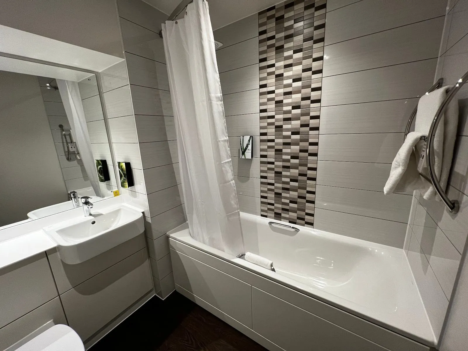 A bathroom at Premier Inn London Paddington (Paddington Basin) with a bath and shower curtain, a strip of grey and brown mosaic tiles, a basin set in a white unit and a curved towel rail