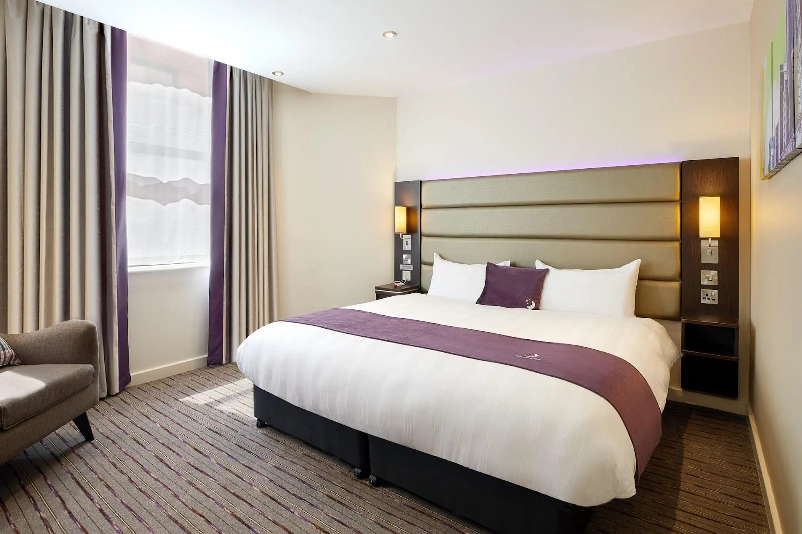 A Premier Inn Kensington (Olympia) room with a wide bed under a padded headboard lit purple from above, a purple runner, a grey armchair and purple-edged curtains at a tall window