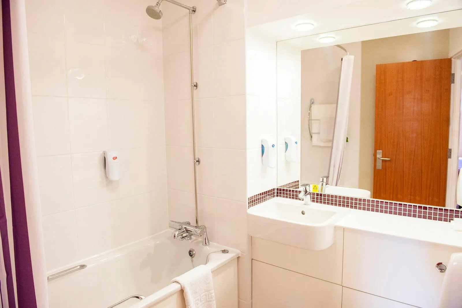 A white-tiled bathroom at Premier Inn London Victoria with a bath and overhead shower, a shower curtain, and a basin set in a white vanity under a wide mirror