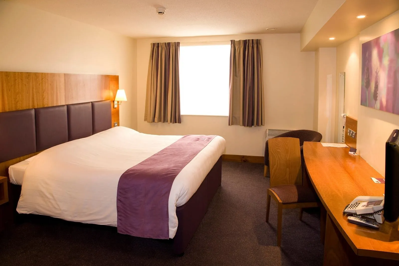 A double room at Premier Inn London Wembley Park with a purple panelled headboard on a pale wood backboard, striped curtains at a wide window and a curved wooden desk