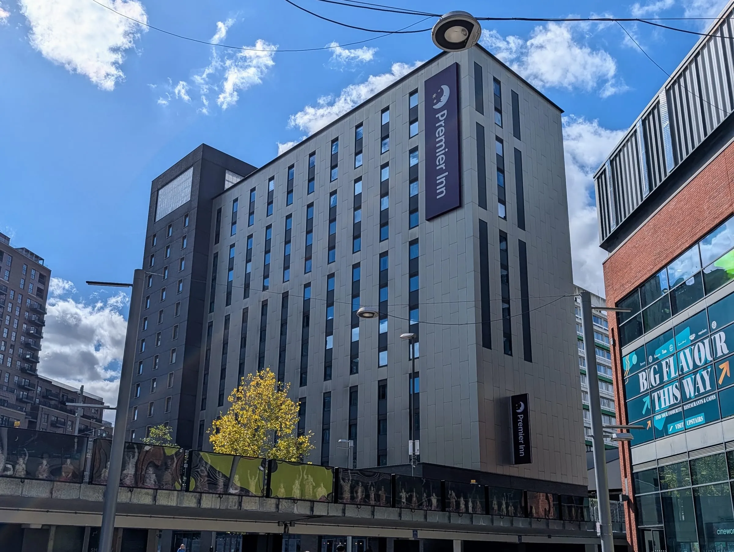 Premier Inn London Wembley Stadium, a tall grey block with purple Premier Inn signs, beside the London Designer Outlet under a blue sky