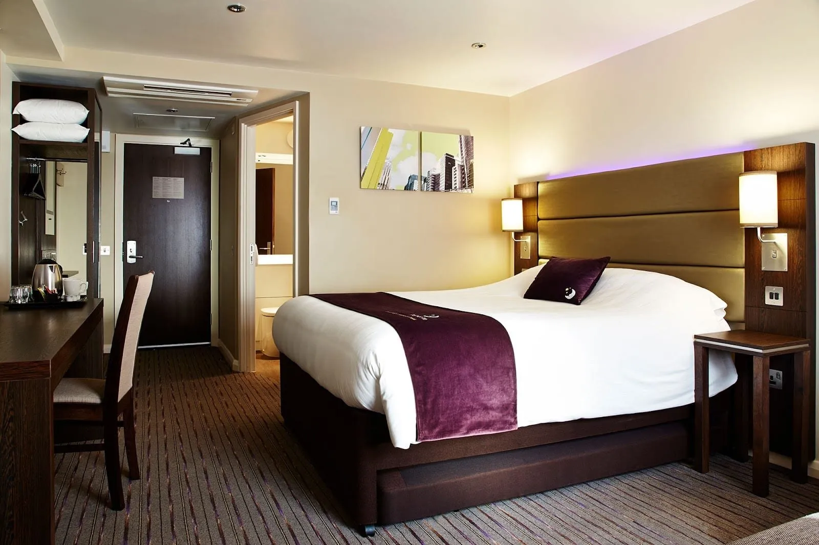 A double room at Premier Inn London Wembley Stadium with a gold padded headboard lit purple from above, a purple bed runner and a dark wood desk by the door