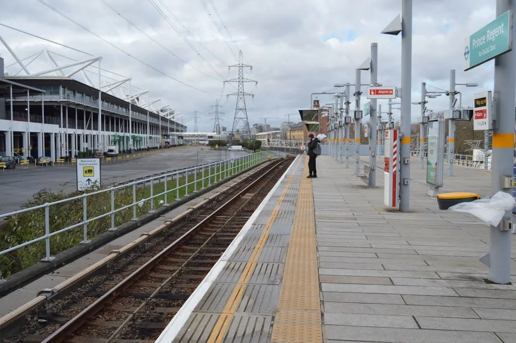 The Prince Regent DLR platform sign, with ExCeL's building running alongside the tracks and electricity pylons on the skyline