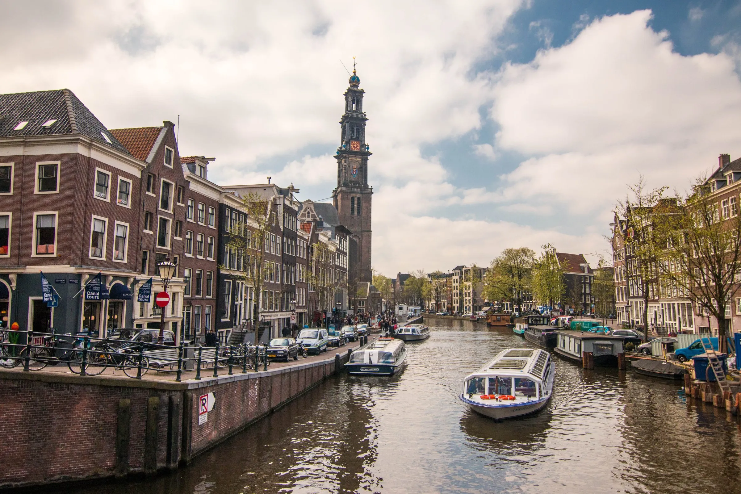 A canal cruise boat passes gabled Amsterdam houses on the Prinsengracht, with the Westerkerk's bell tower rising behind the rooftops on a bright afternoon
