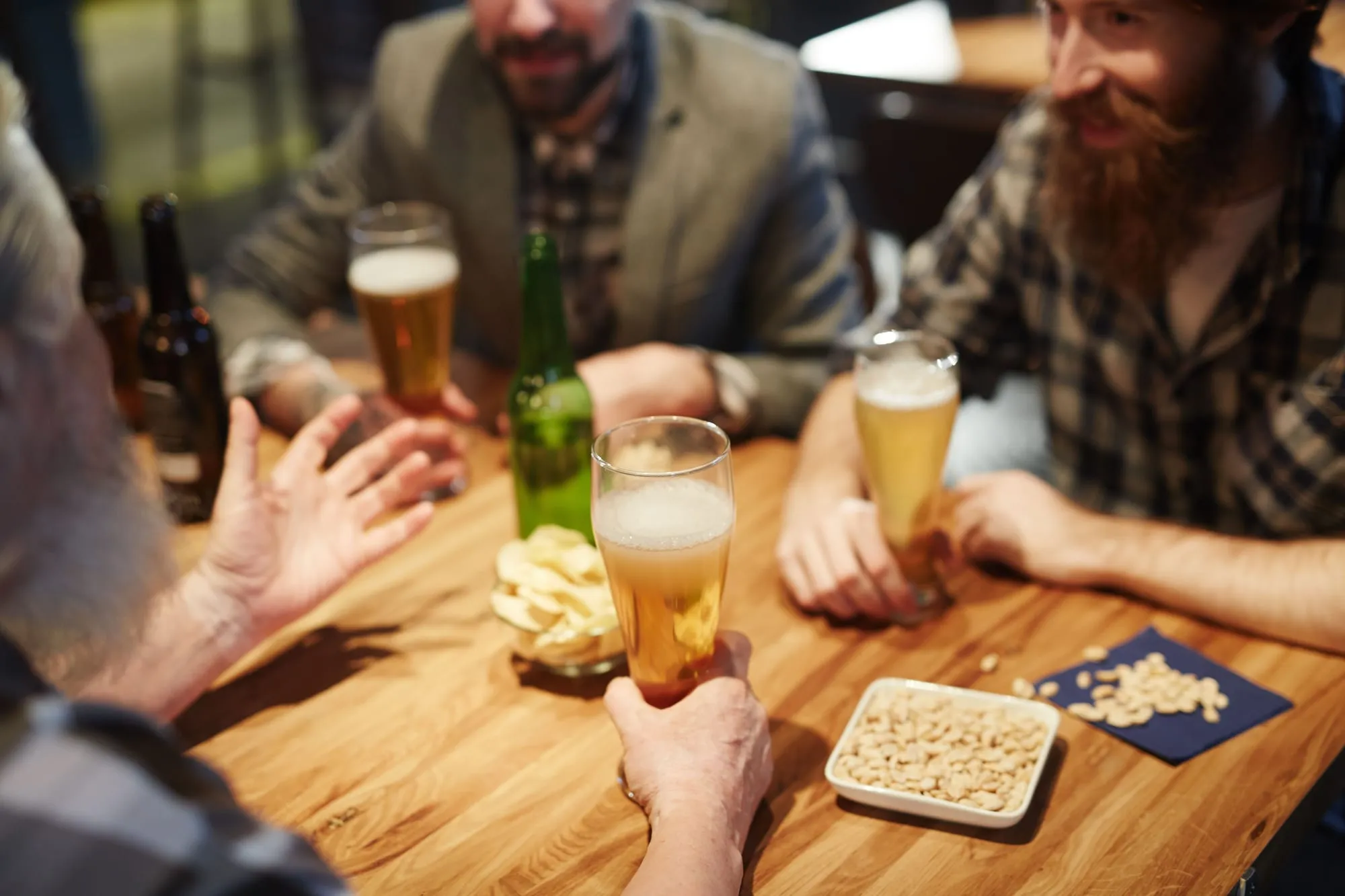 A group of men around a wooden pub table with glasses of beer, bottles, crisps and a dish of nuts between them