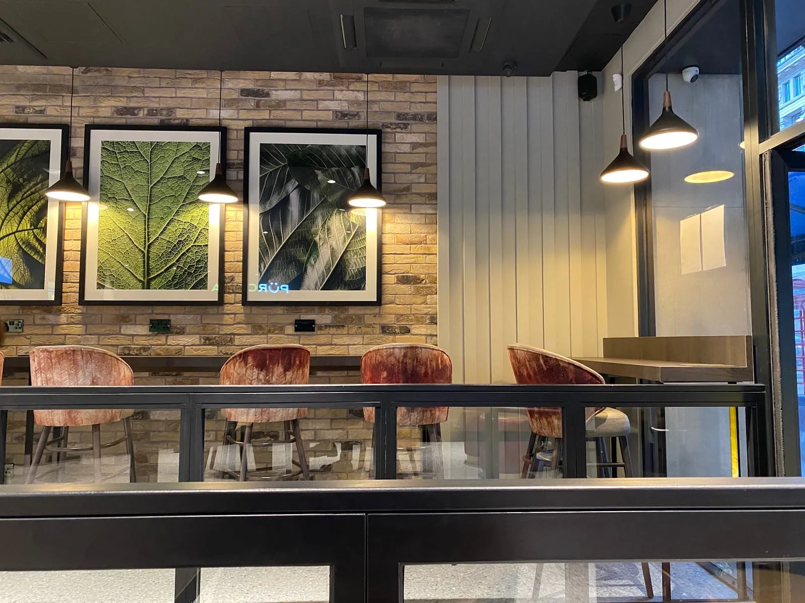 Velvet chairs and a long counter table inside Pürcha, with framed leaf prints on a brick wall and pendant lamps overhead