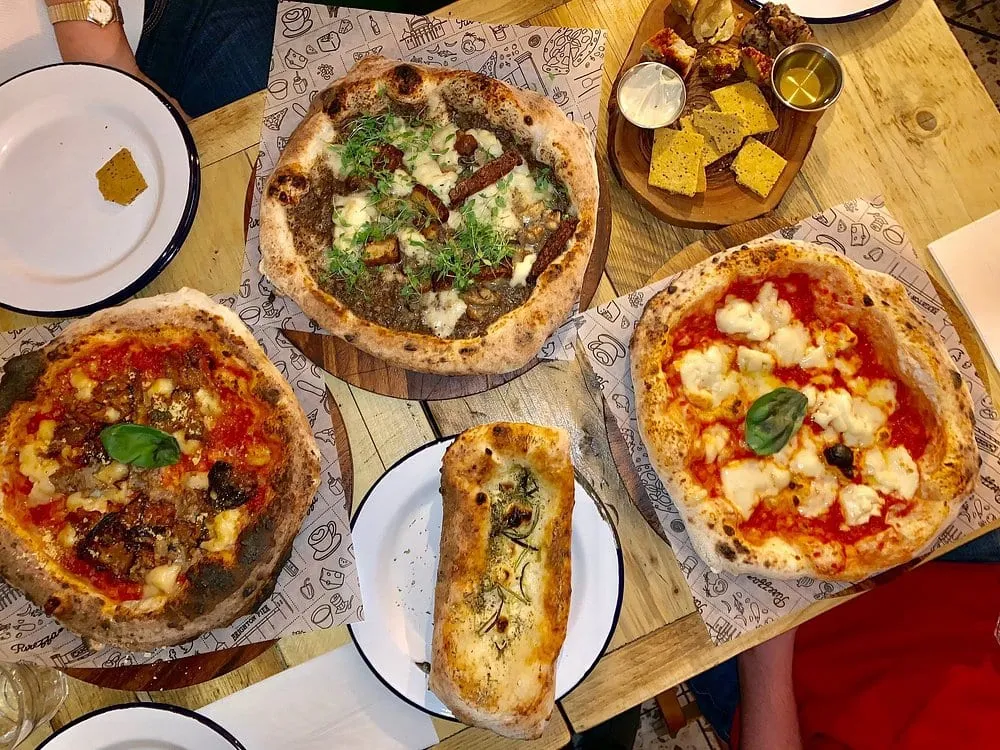 Three vegan pizzas seen from above on Purezza-branded paper, with a board of vegan cheeses and dips and a rosemary focaccia stick between them