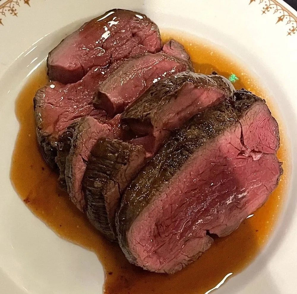 Slices of rare beef resting in a pool of dark jus on a white plate with a gold-patterned rim