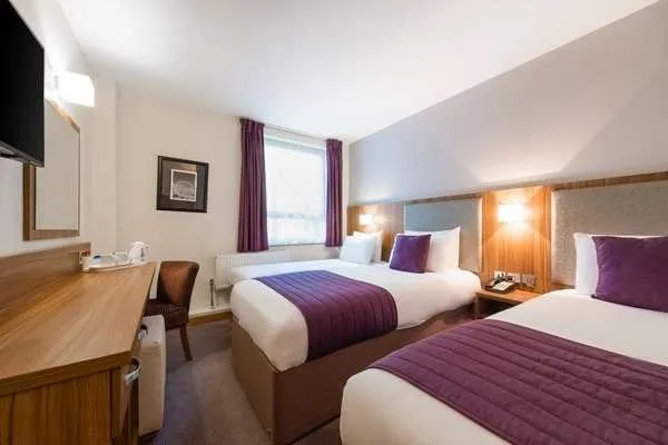A twin room at the Quality Hotel Hampstead with two single beds under purple throws and cushions, grey padded headboards, a window with purple curtains and a long wooden desk under a wall-mounted TV