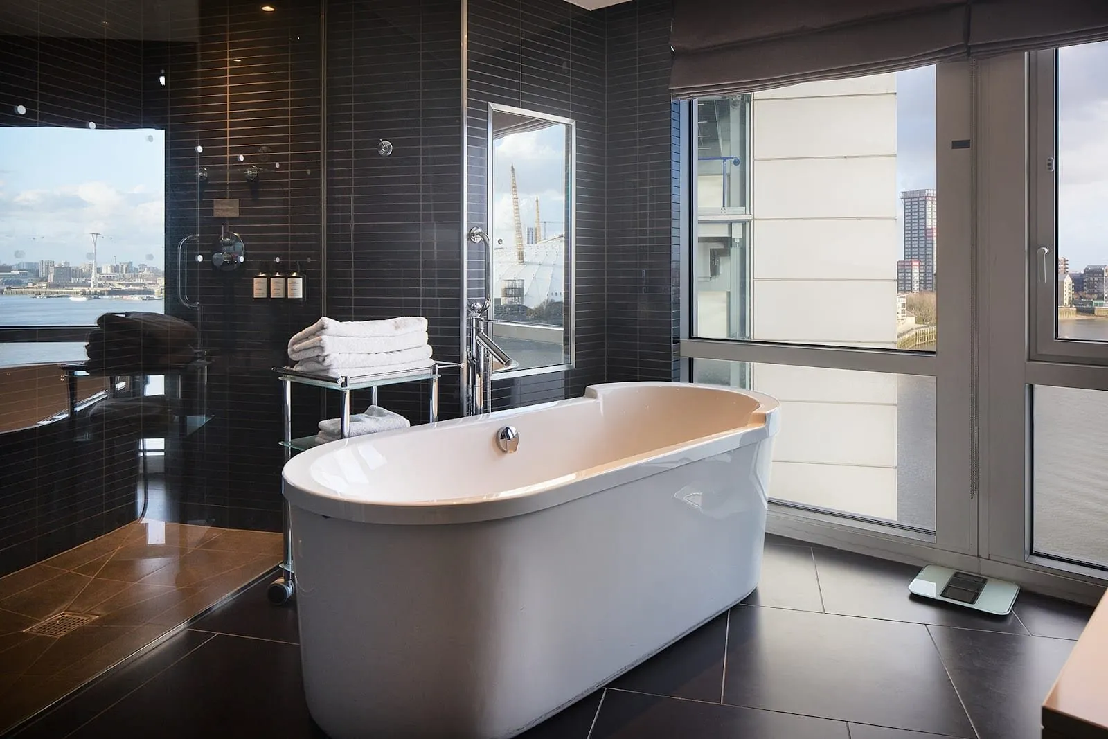 A bathroom at the Radisson Blu Hotel, London Canary Wharf East with a freestanding bath beside full-height windows over the Thames, a glass walk-in shower in dark tiles, and The O2 and the cable car outside