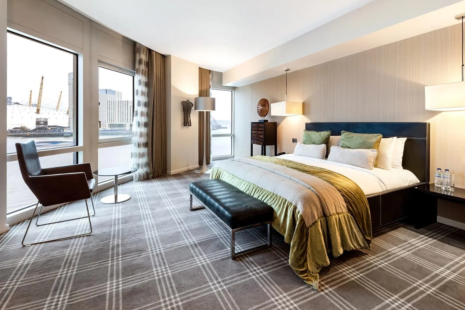 A corner room at the Radisson Blu Hotel, London Canary Wharf East with a king bed under an olive throw, a leather bench at its foot, and windows on two sides looking across the Thames to The O2