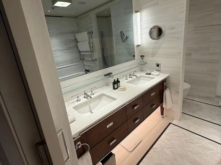 A bathroom at Raffles London at The OWO with twin basins in a white marble counter over dark wood drawers, a wide mirror and a herringbone marble floor