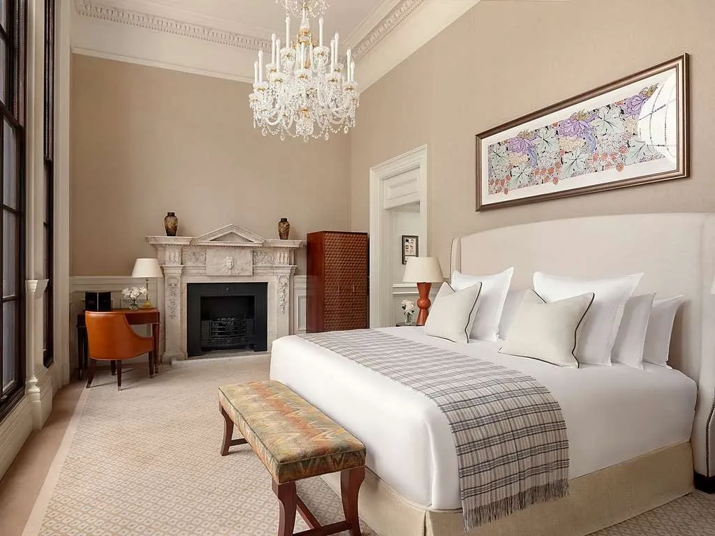 A high-ceilinged room at Raffles London at The OWO with a crystal chandelier, a carved marble fireplace, a large bed with a checked throw, a long floral print and tall windows