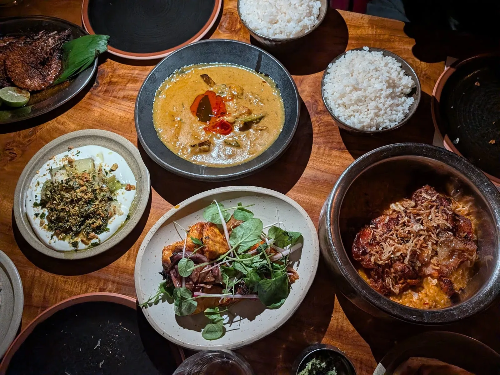 An overhead table of shared dishes: a whole fish crusted dark brown on a banana leaf with lime, a wide bowl of pale coconut curry topped with red chillies, two bowls of white rice, a plate of charred pieces with red onion and watercress, and a metal pot of curry under a heap of crisp fried shallots