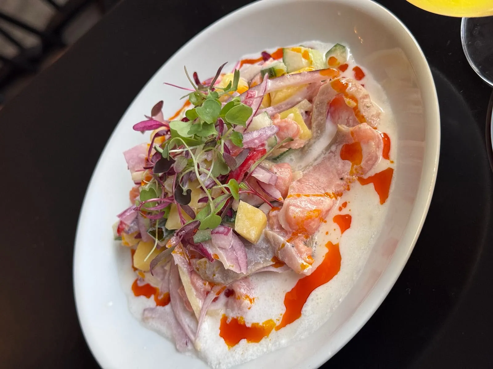 An oval plate of kinilaw: slices of raw pink fish in a milky dressing with red onion, cucumber, diced mango, micro herbs and a drizzle of orange chilli oil