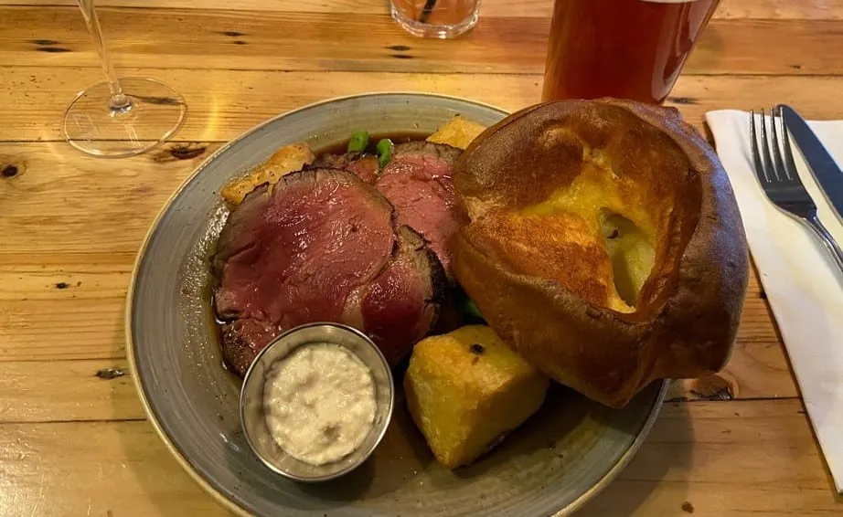A roast beef dinner at The Red Lion and Sun: pink slices of beef in gravy with roast potatoes, a tall Yorkshire pudding and a pot of horseradish, on a pub table beside a pint