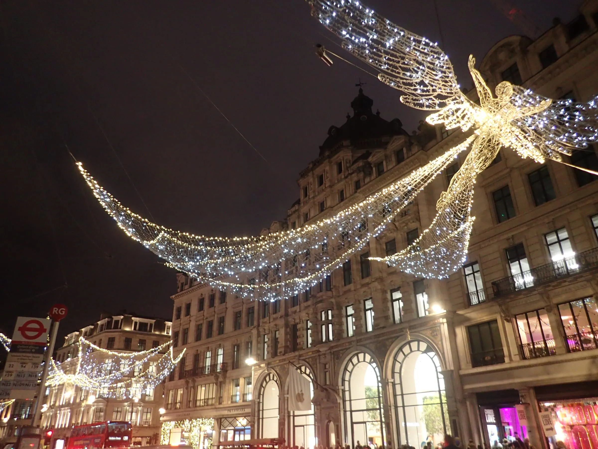 A Spirit of Christmas angel above Regent Street, its lit wings spanning the road between strings of white lights, with the Apple store's arched windows below