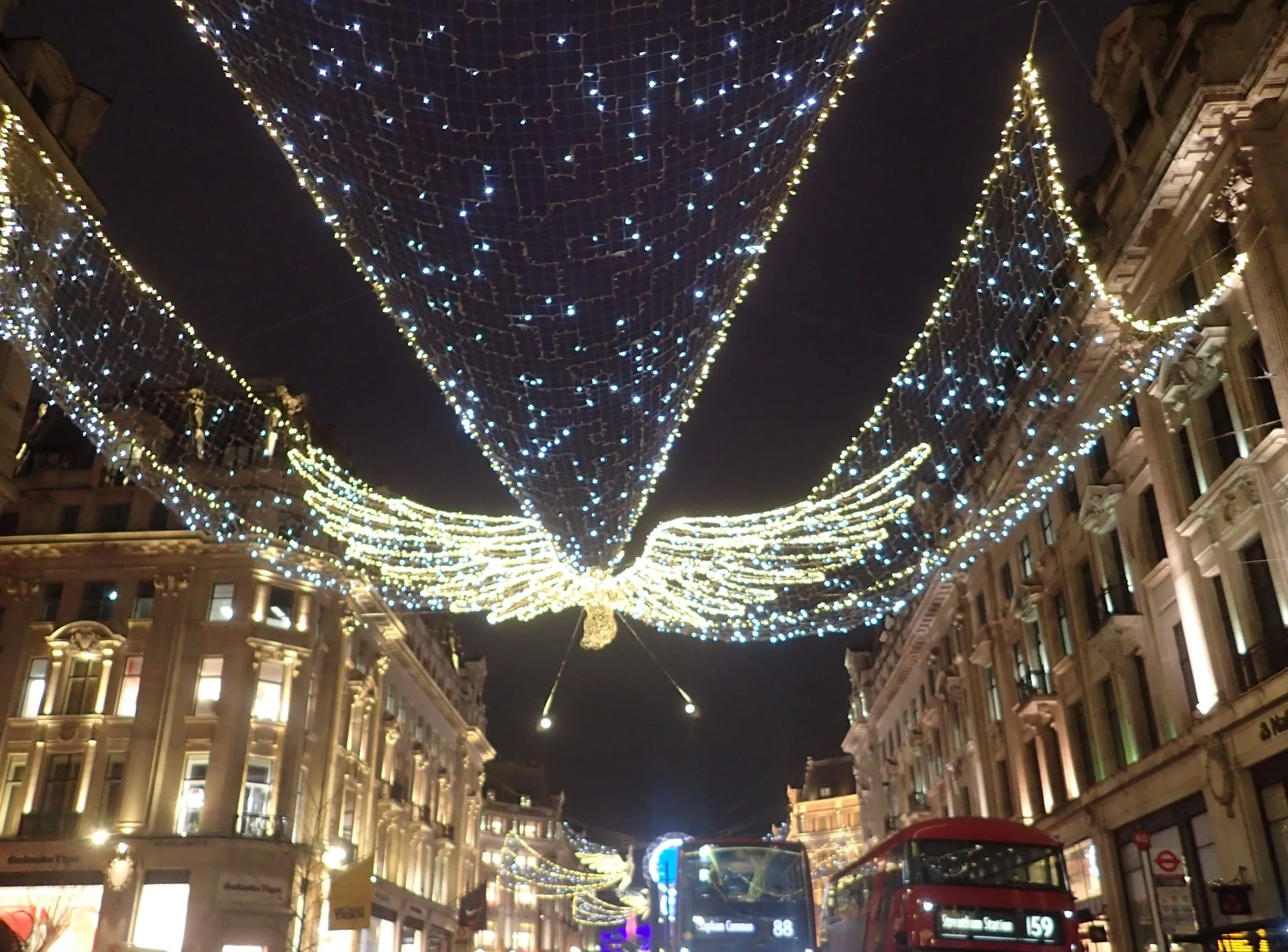 One of the Spirit of Christmas angels, wings spread under a canopy of white lights, above Regent Street at night with two red buses below