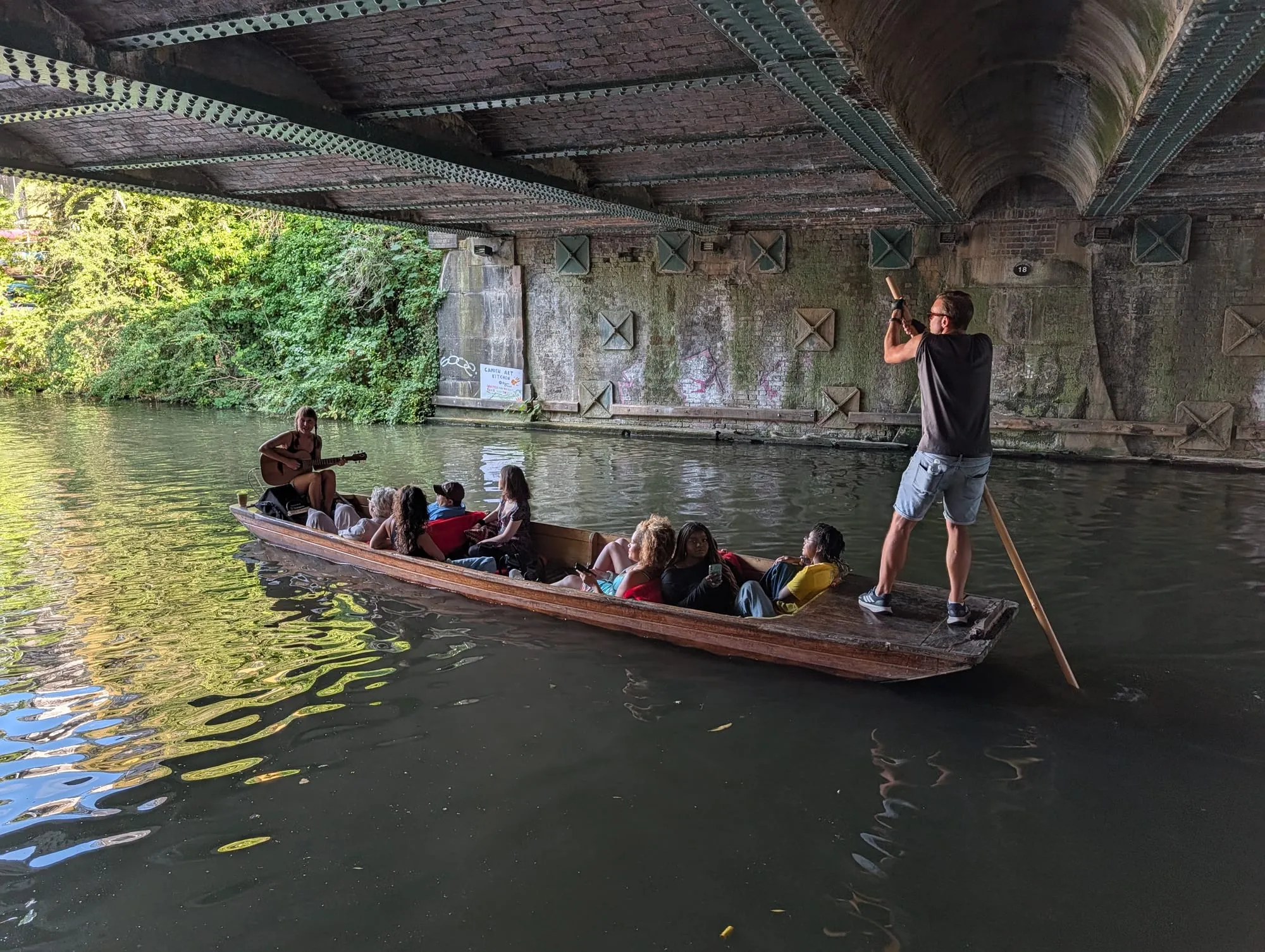 A wooden punt on the Regent's Canal under a bridge, with a guitarist playing to the passengers aboard