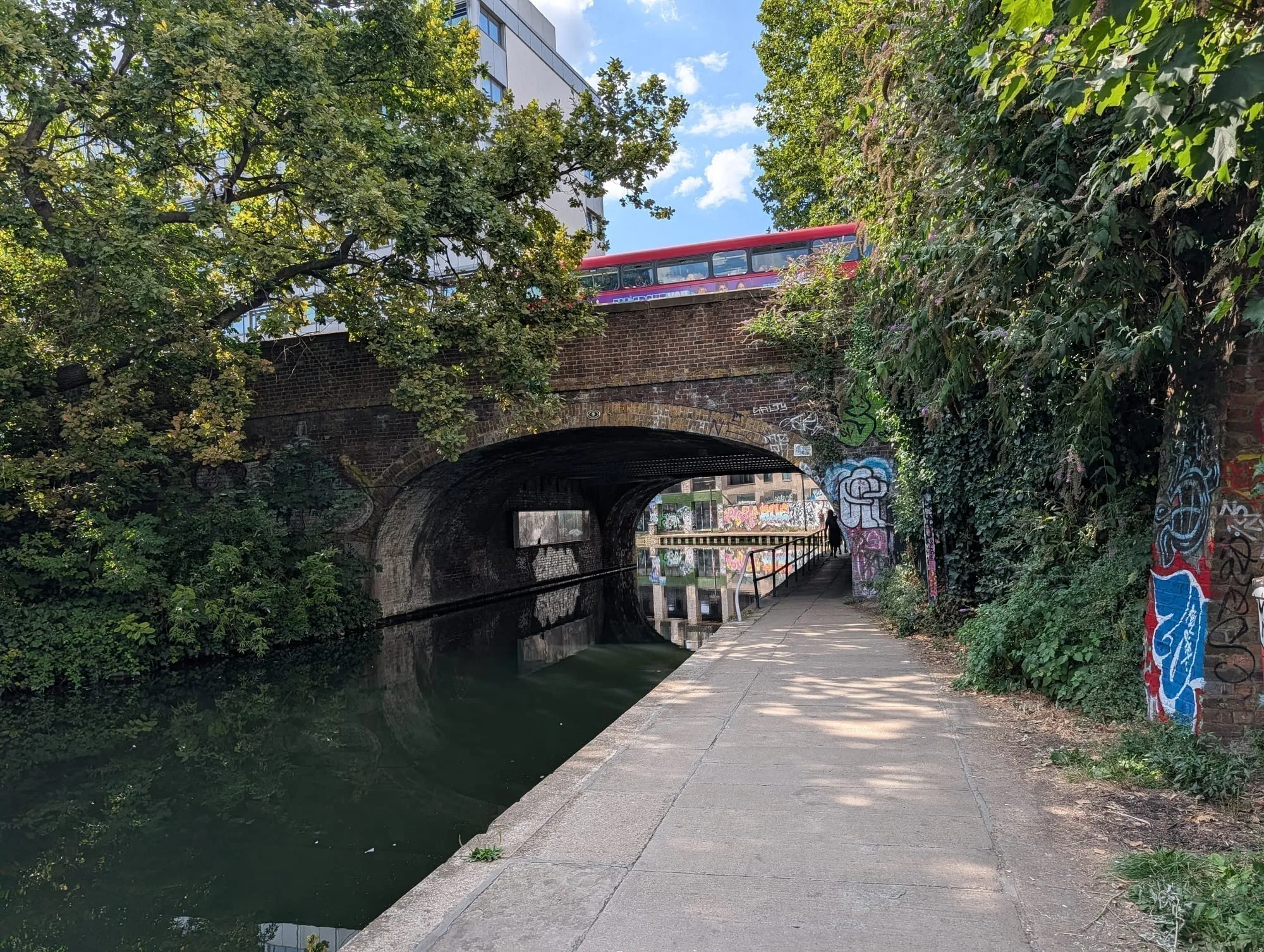 A brick canal bridge over the Regent's Canal with a red double-decker bus crossing above it, graffiti on the abutments and the towpath running under the arch