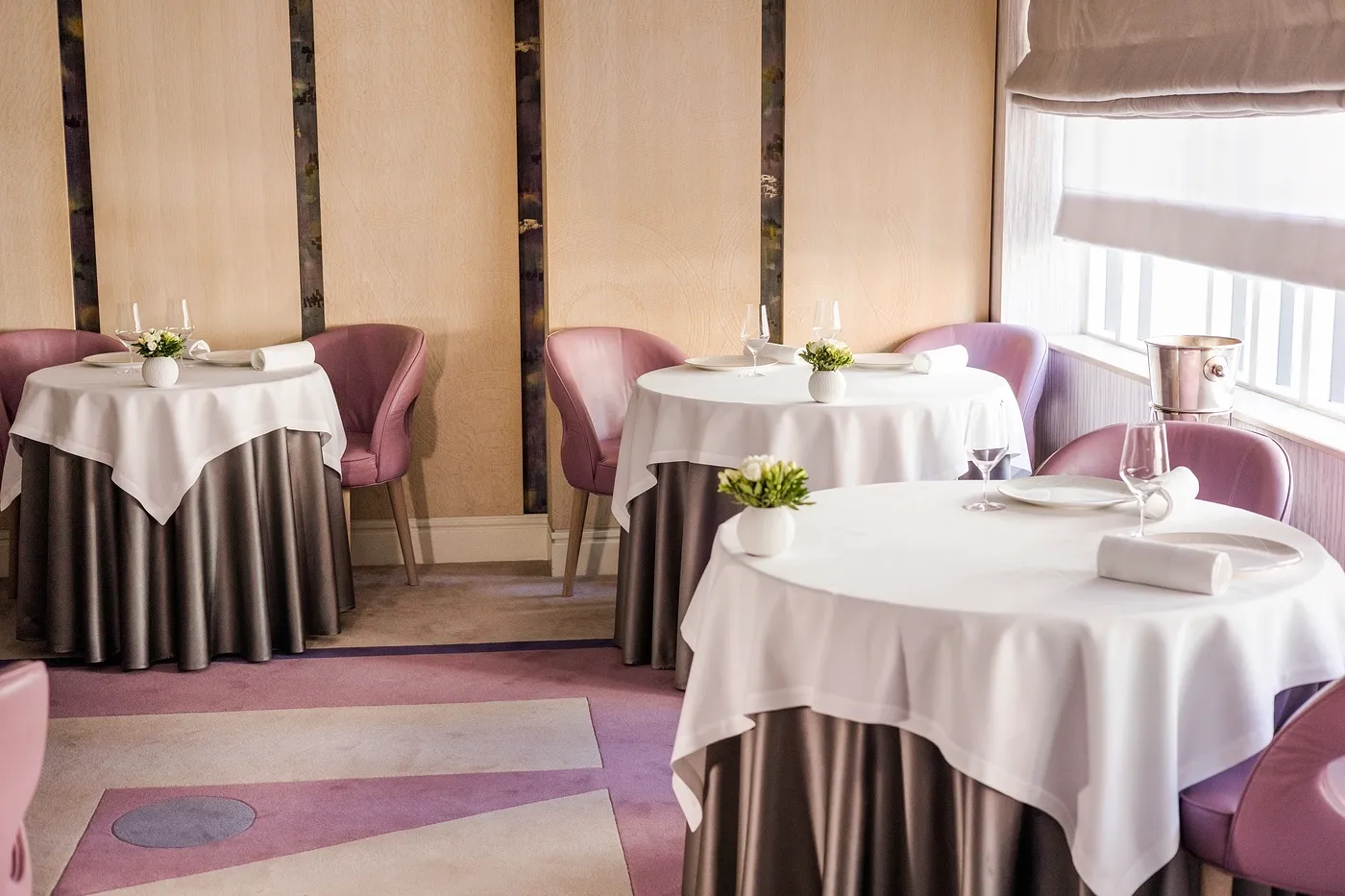 A small dining room at Restaurant Gordon Ramsay, with pink-grey upholstered chairs and round tables laid for service