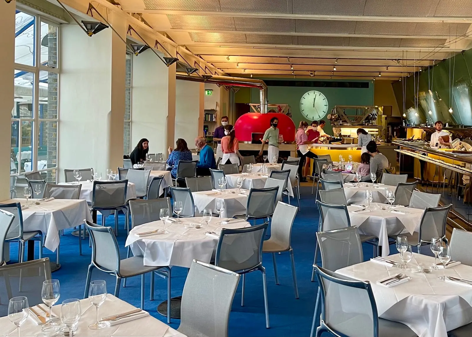 The River Café dining room: white-clothed tables on a blue floor, with an open kitchen along the far wall, a domed red wood-fired oven and a large wall clock above the pass