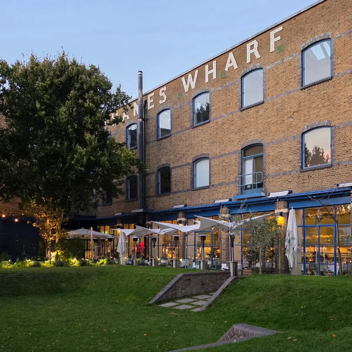 The River Café's lit terrace on the Thames at dusk, beneath the Thames Wharf sign, with tables and patio heaters set outside