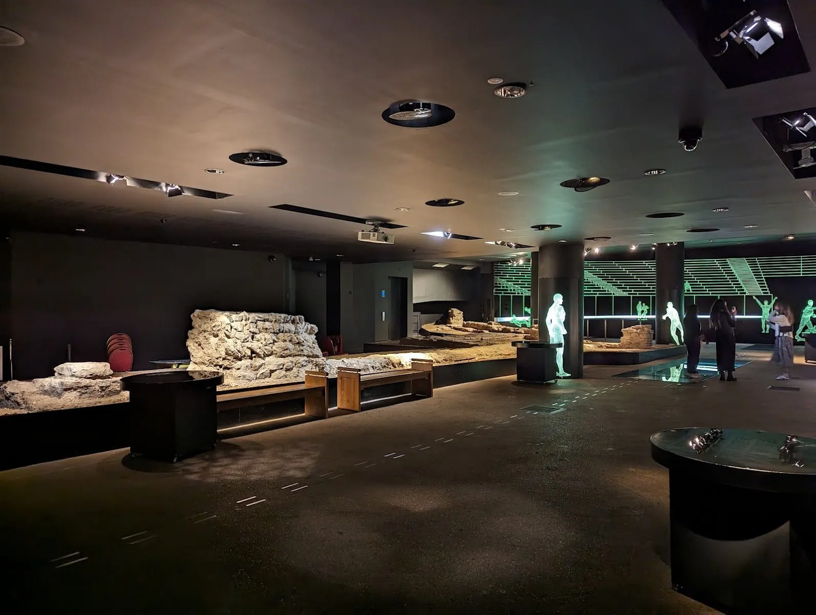 A dark basement gallery at the Guildhall Art Gallery, with a low stretch of Roman stone wall lit from above and green outlines of spectators on the far wall