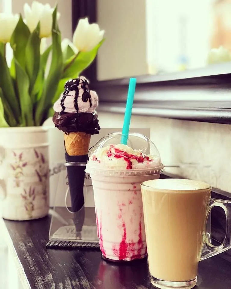 A cone with a dark chocolate-dipped shell and pink gelato, beside a raspberry milkshake and a latte on a dark windowsill with white tulips
