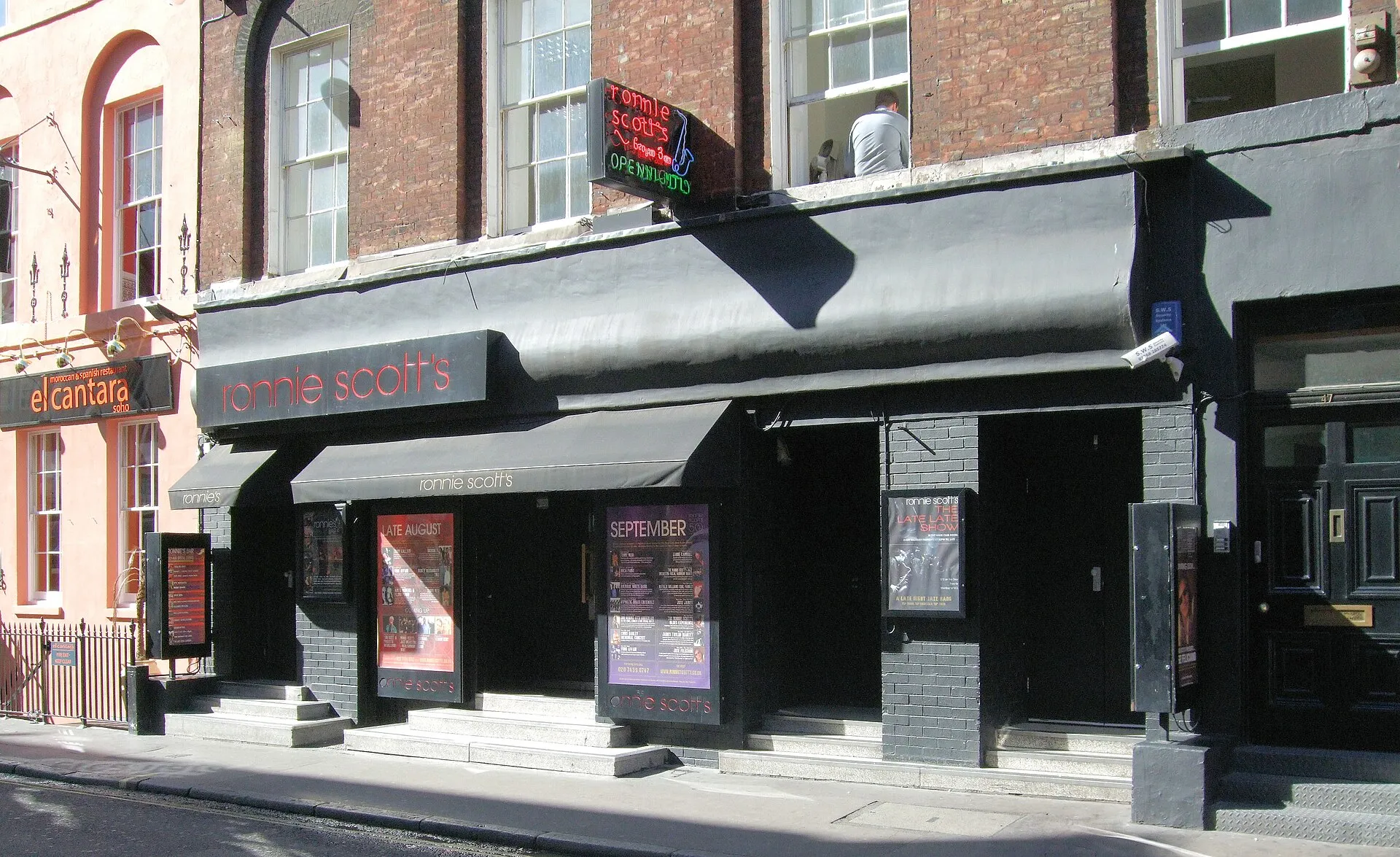 The frontage of Ronnie Scott's on Frith Street in Soho, its neon sign above a black awning and the month's bills in lit cases beside the door