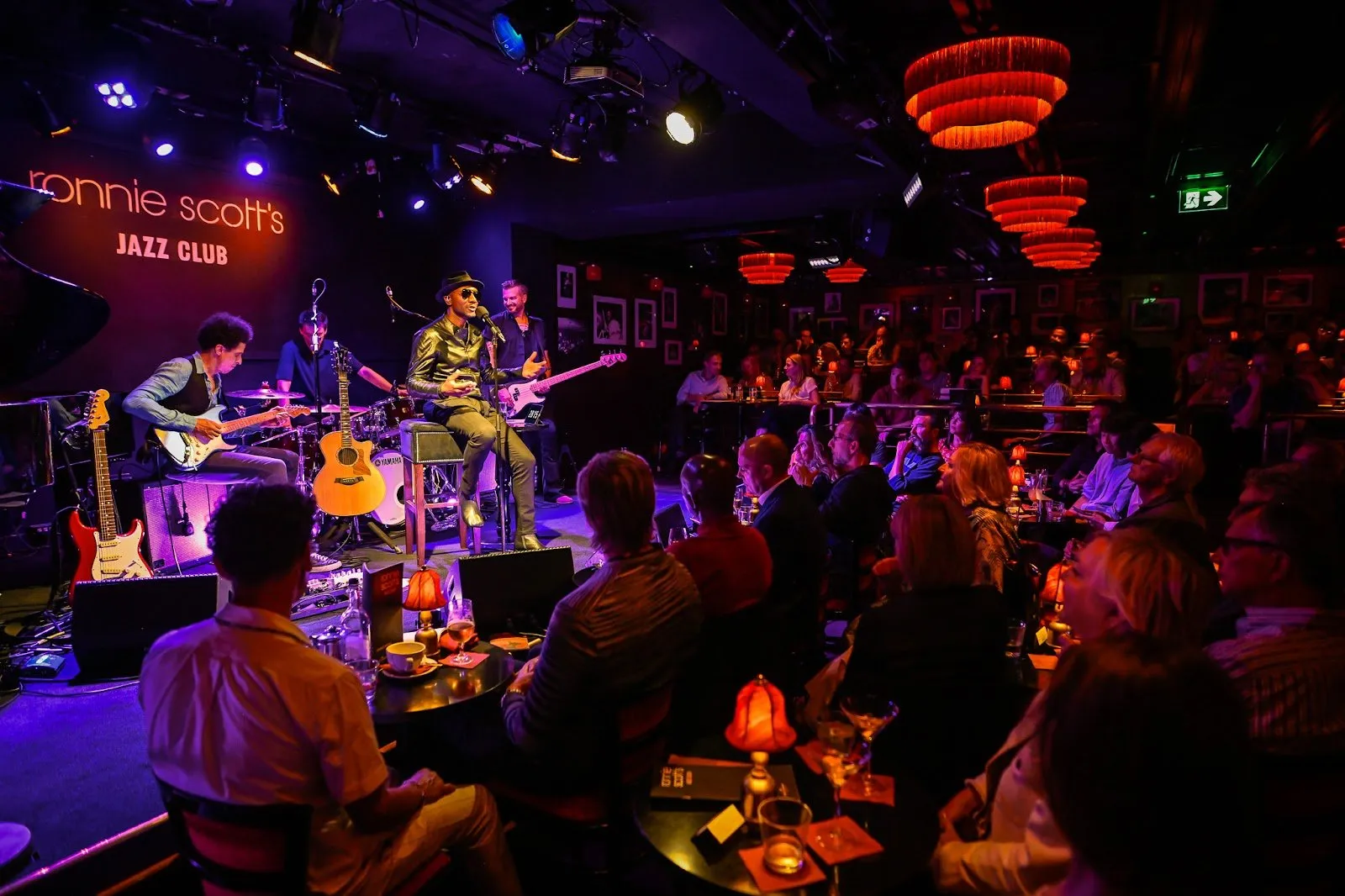 A band plays on the Ronnie Scott's stage under the club's neon sign, with the audience at small lamplit tables in front