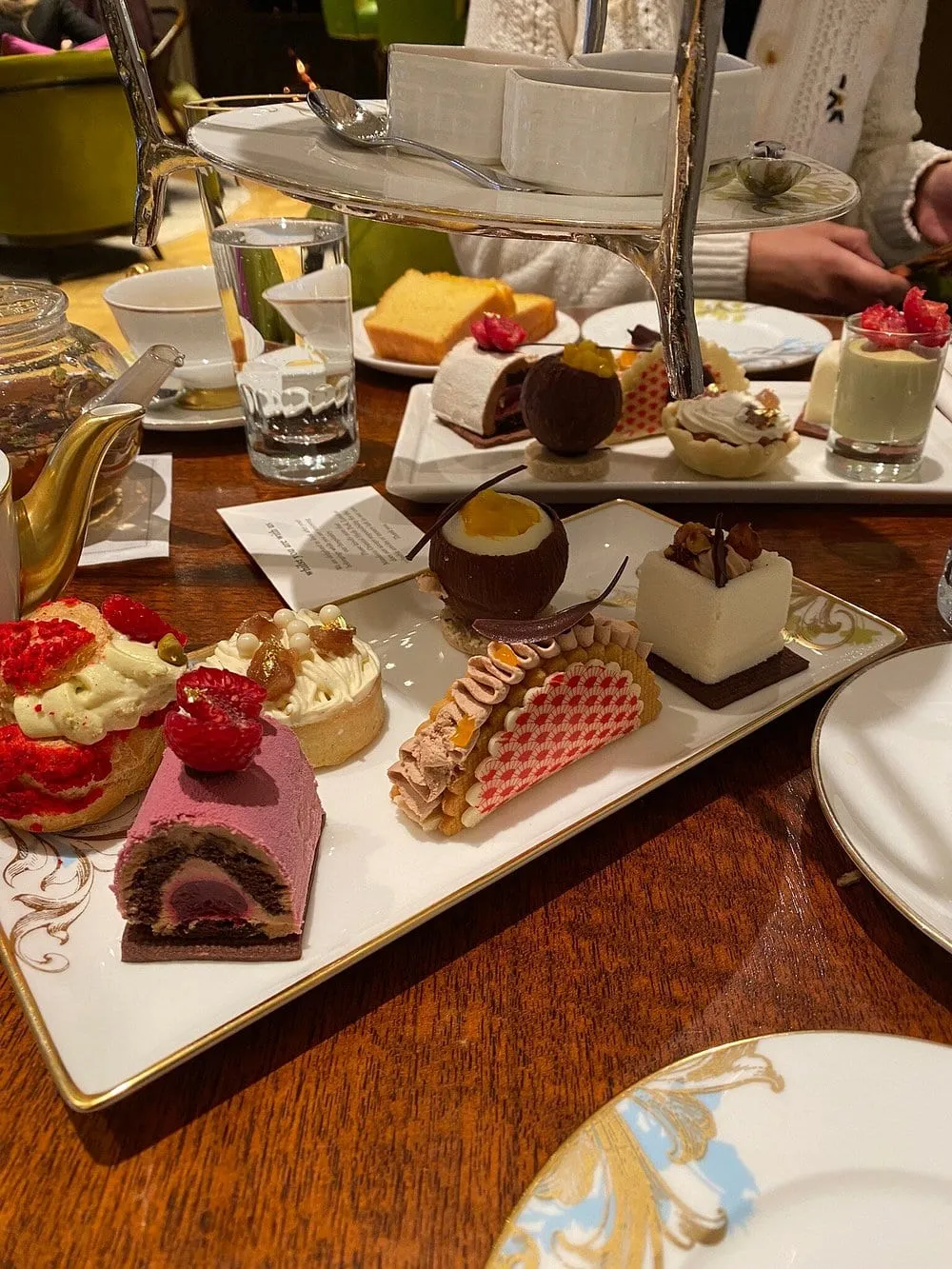 A tiered stand of pastries on gold-rimmed china in the Rosebery Lounge, with a gold teapot and green velvet chairs behind