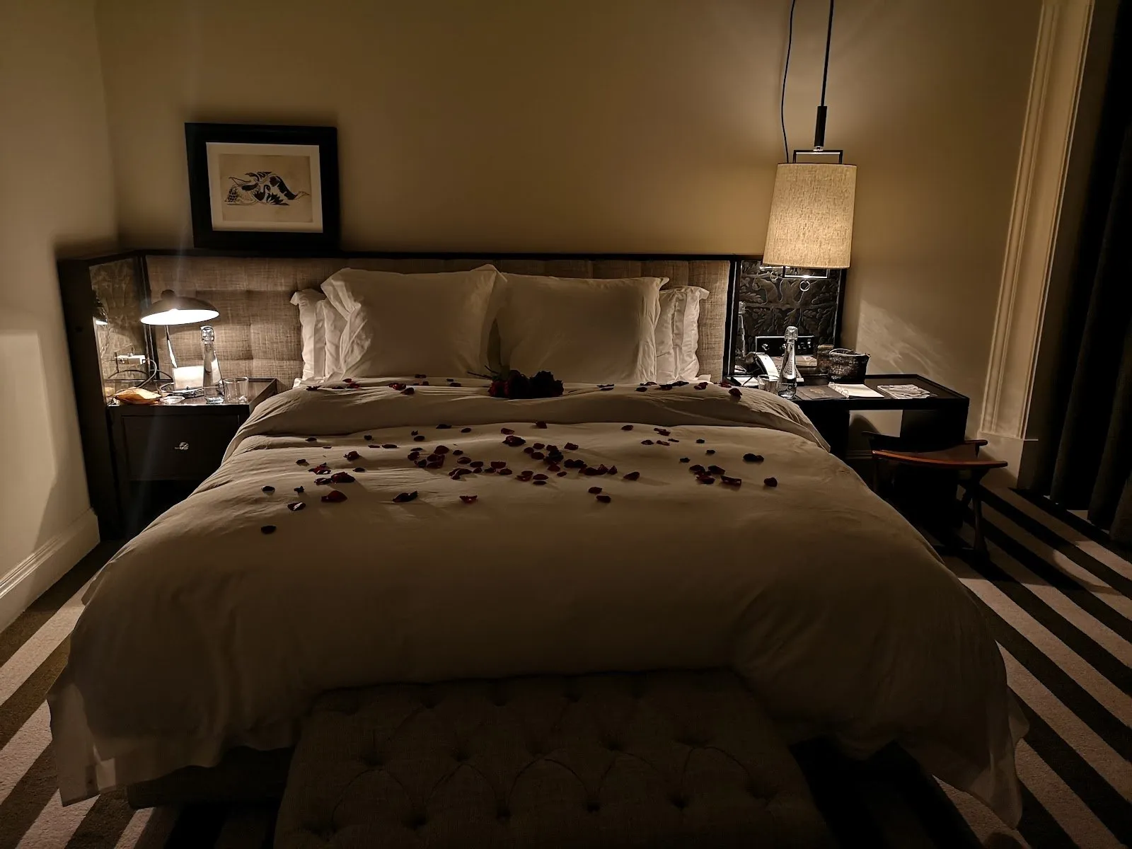 A bed strewn with rose petals at Rosewood London, with a tufted headboard and a striped carpet underfoot