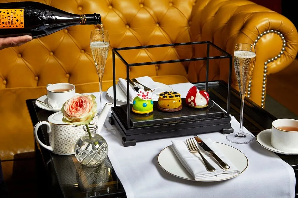 Three brightly coloured art-inspired cakes in a glass display case on a black stand, with champagne being poured against a yellow leather banquette