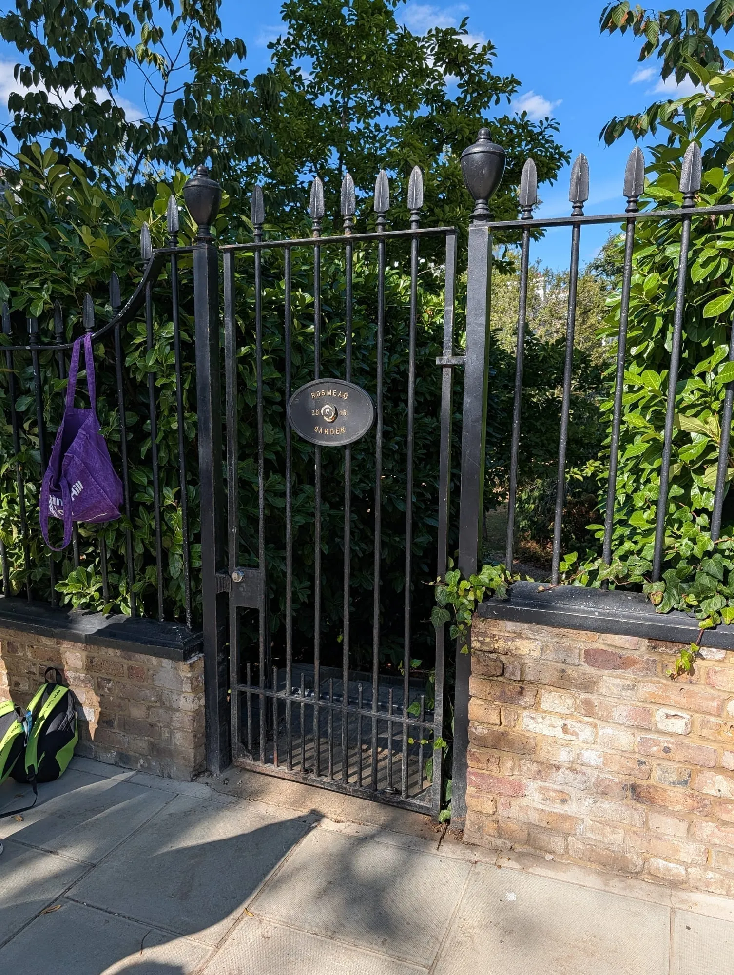 A locked black iron gate to Rosmead Garden with a small oval name plaque, seen from the pavement