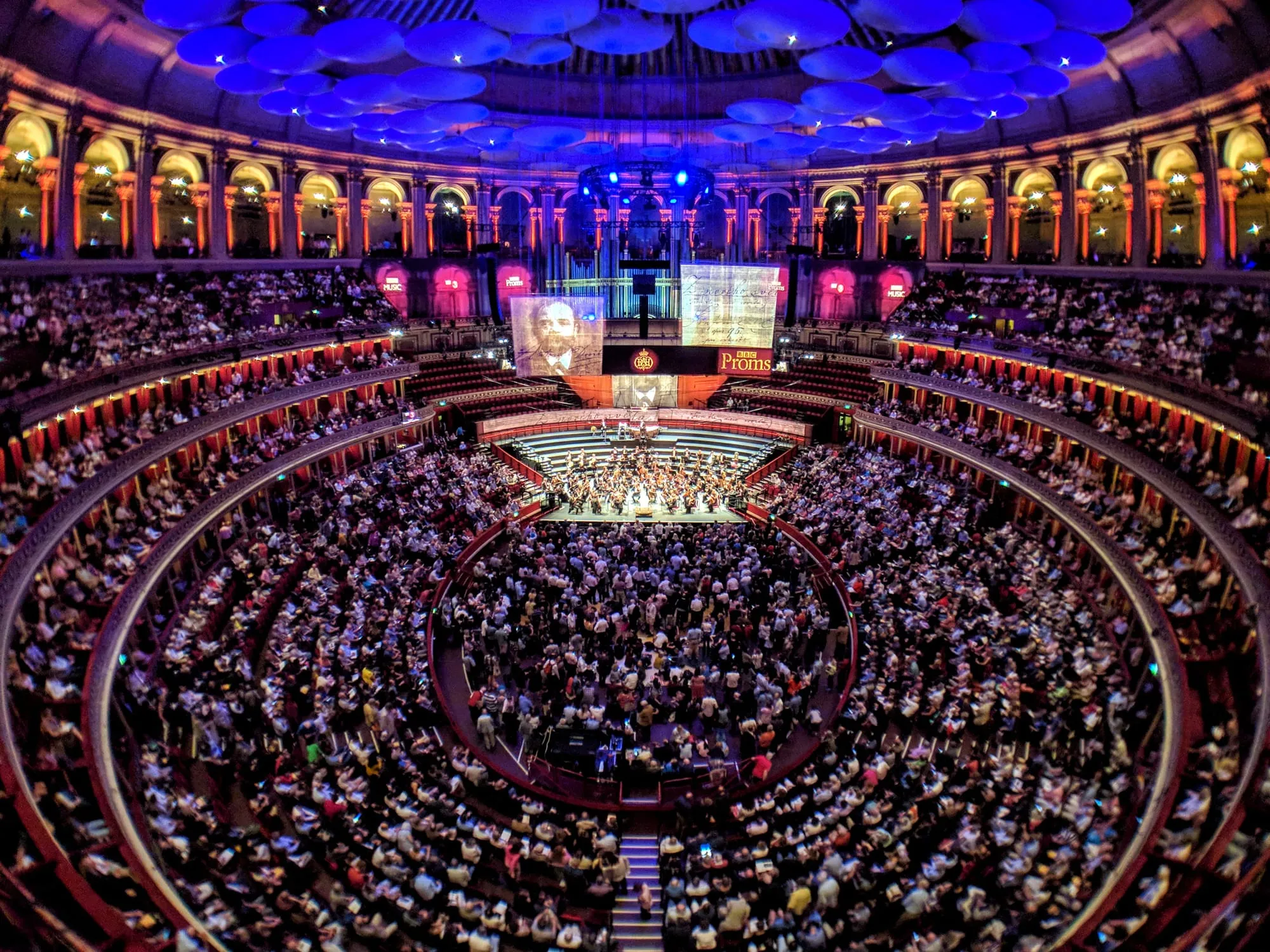 Inside the Royal Albert Hall during a Prom, with the orchestra on stage, the standing Arena in front of it and the circles rising above