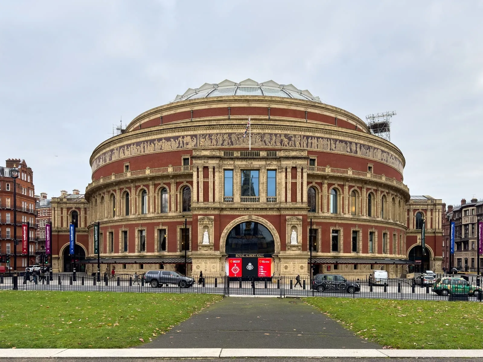 The Royal Albert Hall's red-brick rotunda and terracotta frieze seen across a lawn, under a grey sky