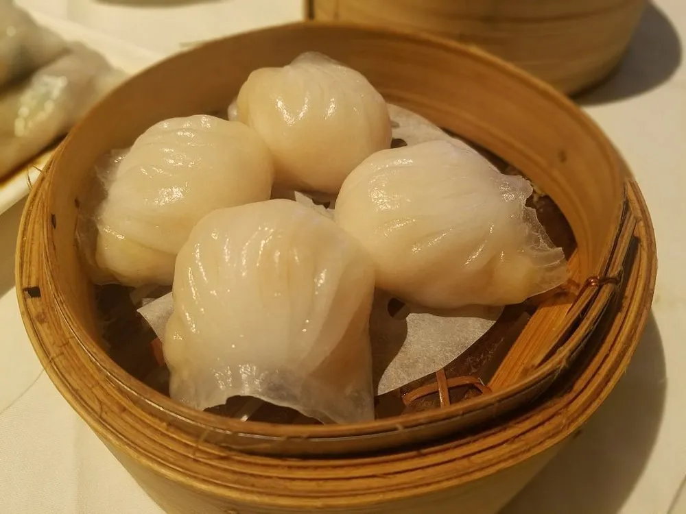 Four har gau prawn dumplings in a bamboo steamer, their pleated translucent wrappers showing the prawn inside