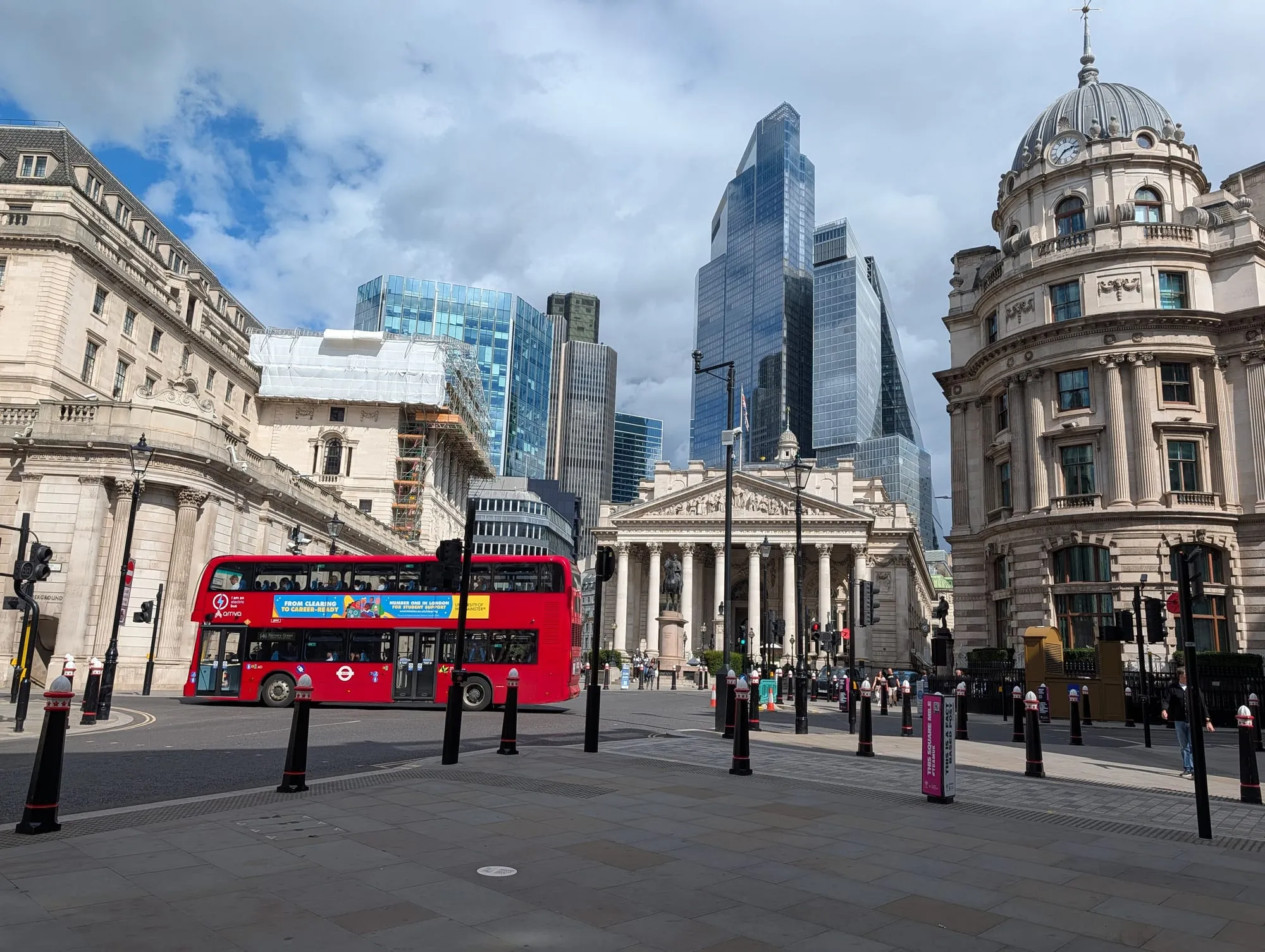 A red bus passing the Royal Exchange at Bank, with the City's towers rising behind it