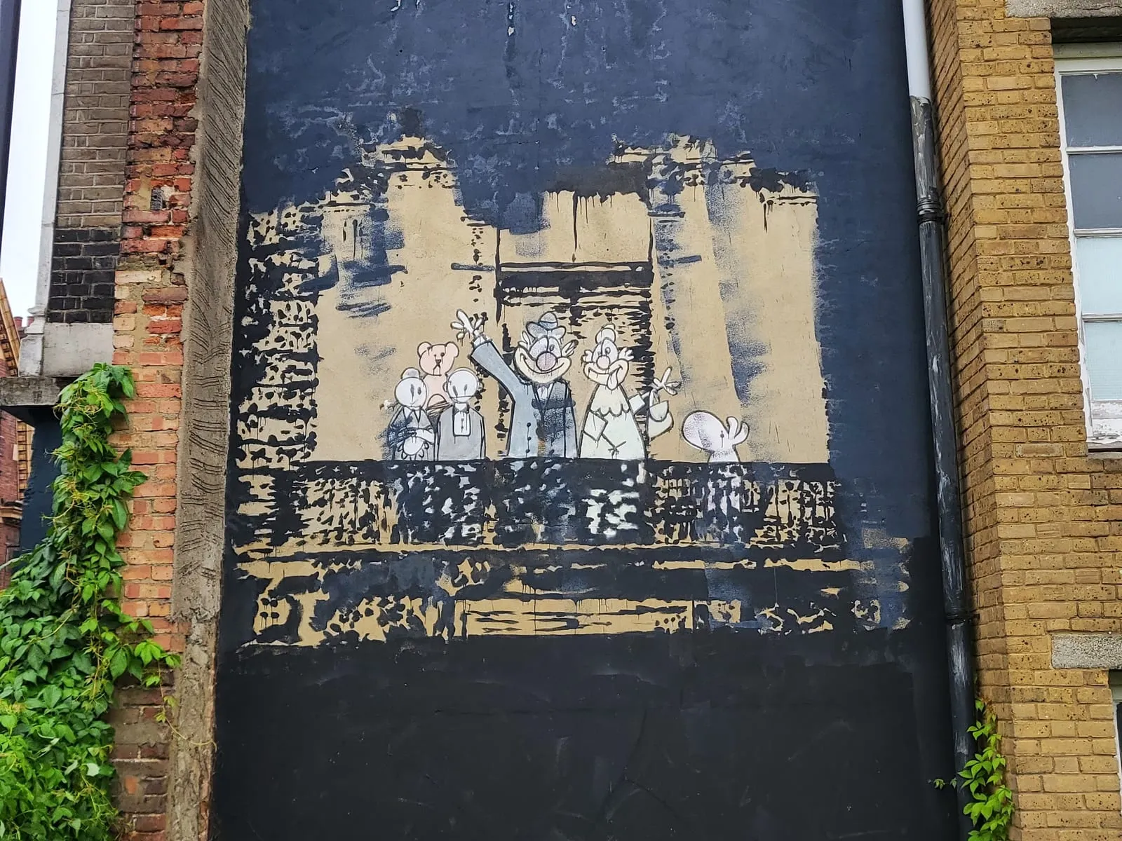 Banksy's Royal Family on the side of a building in Stoke Newington, cartoon royals and a teddy bear waving from a balcony, framed by a patch of beige wall inside a large area of black paint