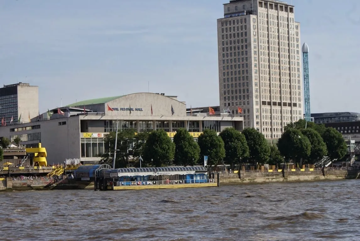 The Royal Festival Hall seen from the Thames, with a pier in front, a row of trees along the embankment and a tall office tower behind