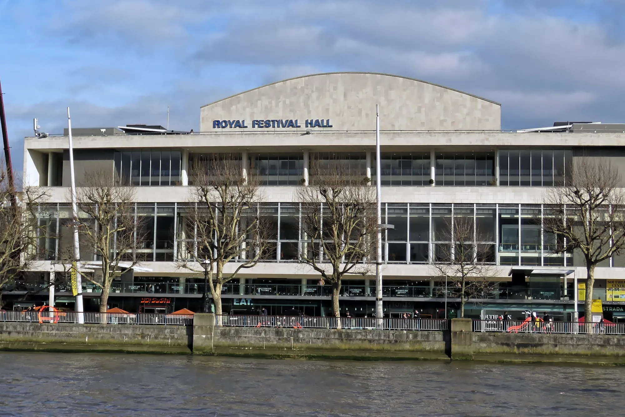 The Royal Festival Hall's stone-clad front on the South Bank, seen across the Thames, with its name in blue letters above the glazed foyers