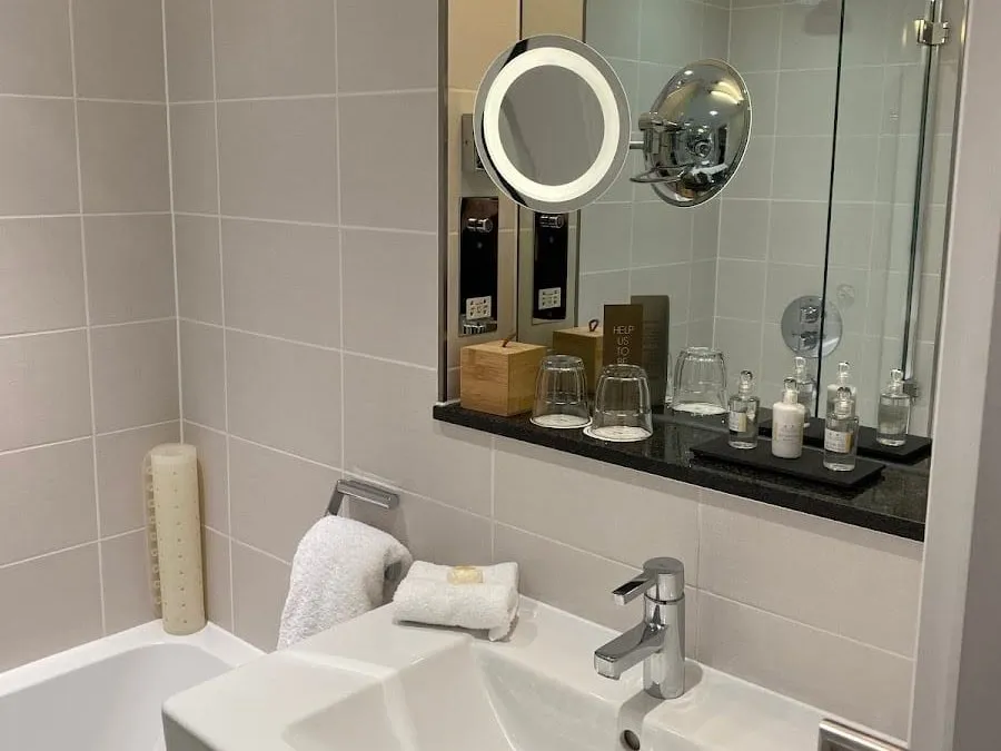 A bath below a basin and a mirror shelf with a lit magnifying mirror, glasses and miniature toiletries at the Royal Garden Hotel