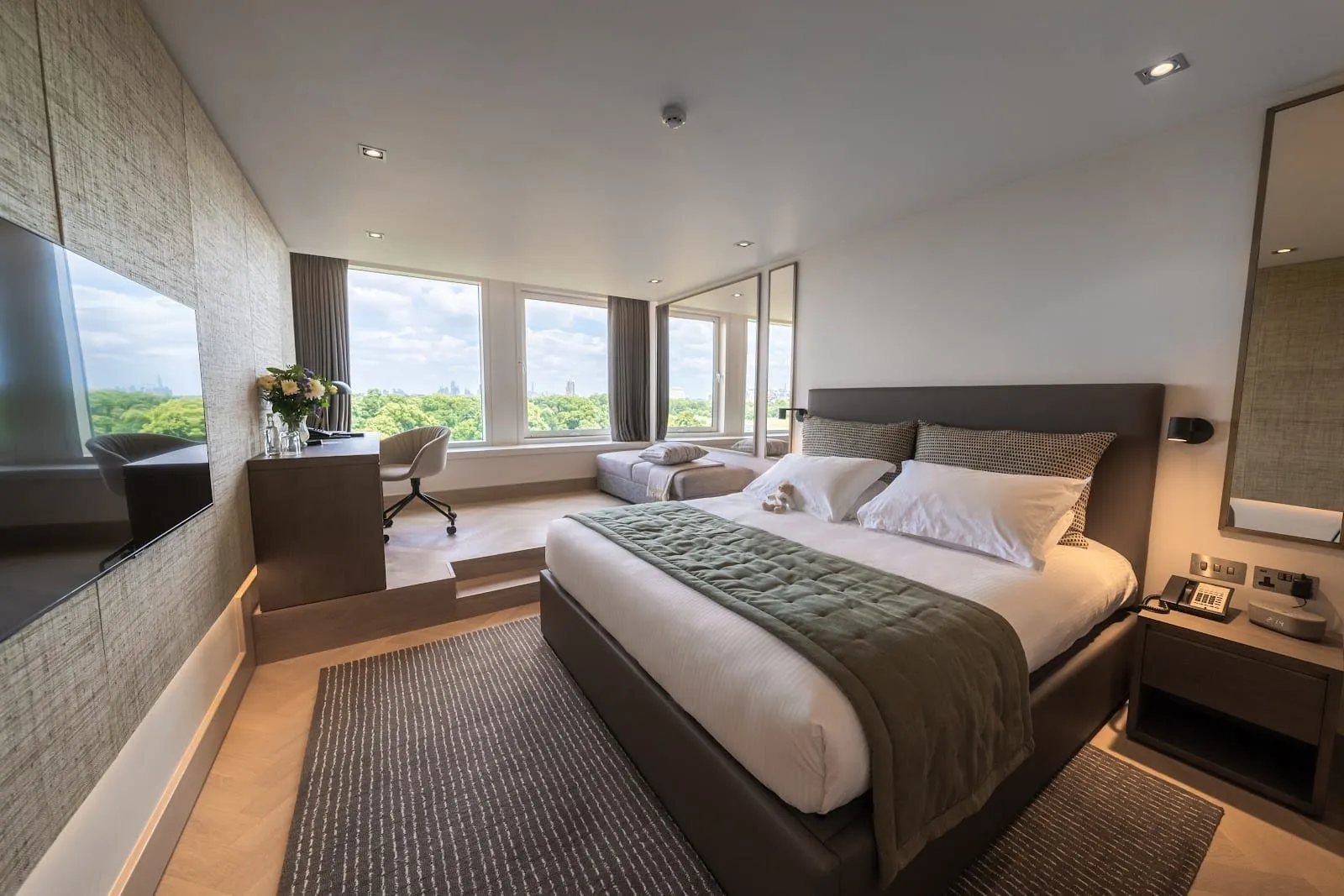 A Royal Garden Hotel bedroom with a dark leather headboard, a green quilted throw and a raised window seat with a desk, looking over the treetops of Kensington Gardens to distant towers