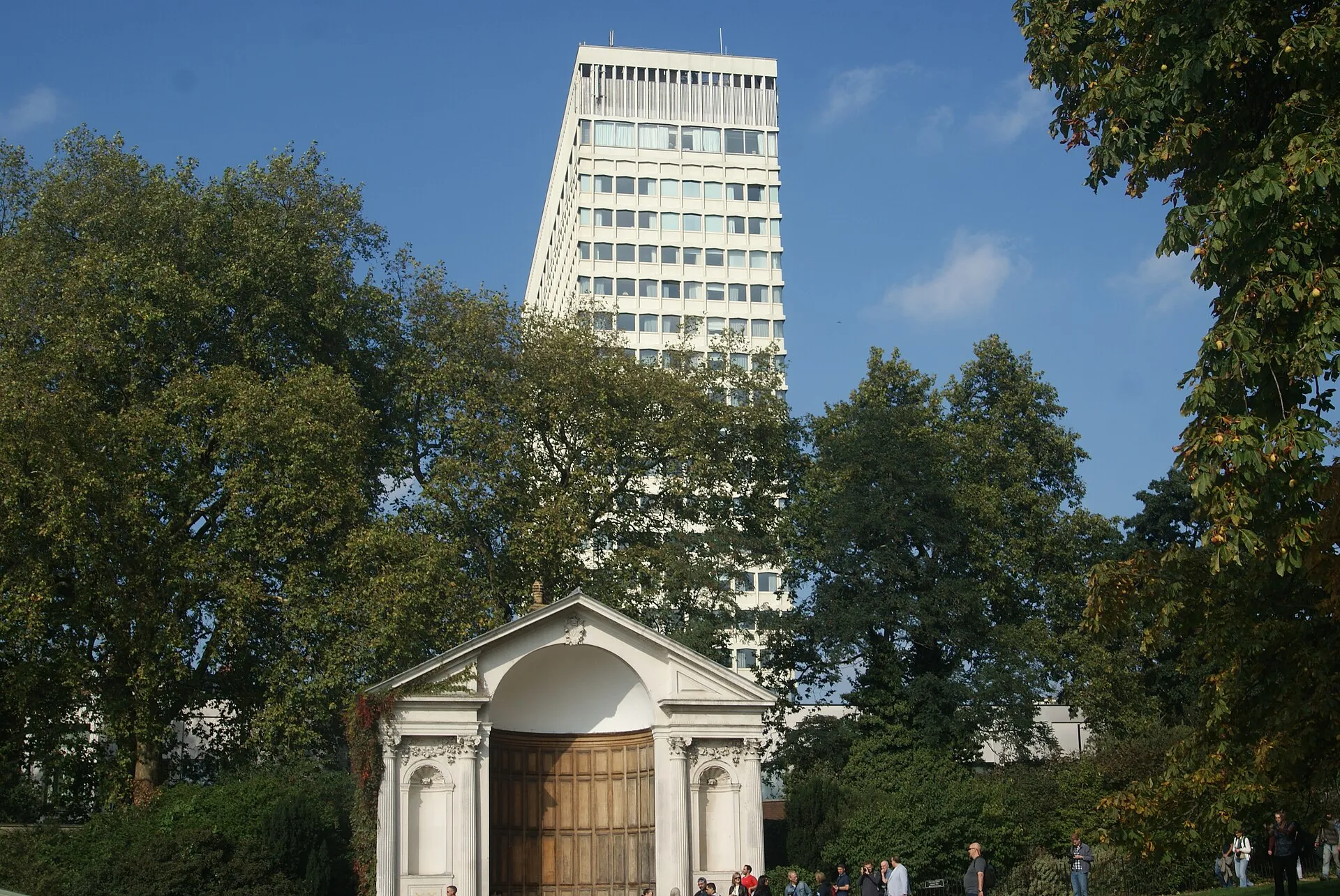 The white tower of the Royal Lancaster rising above the trees behind a stone alcove seat in the Italian Gardens, under a blue sky, with people walking in front